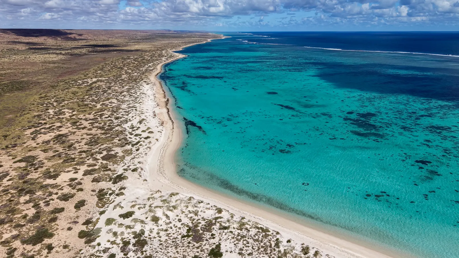 A drone view of a bay in shades of blue, with dunes and coastal bushland.